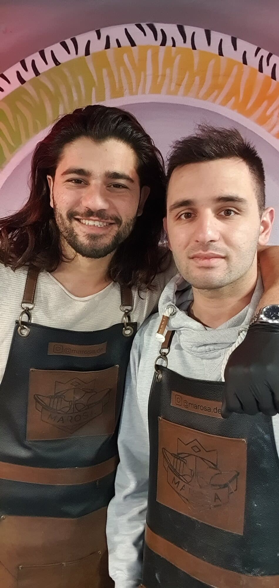Samed und Abdul lachend in Lederschürzen vor dem Wandbild im Laden in Fellbach